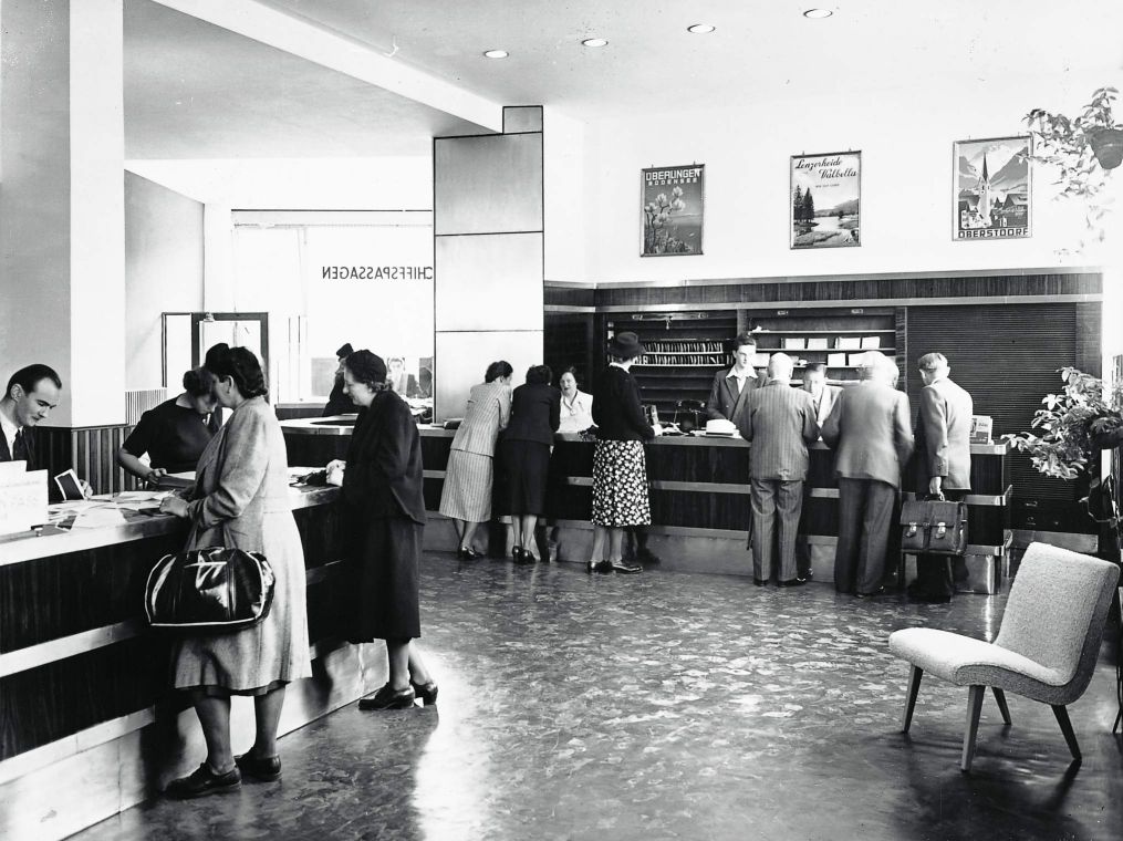 Schwarzweißfoto eines Innenraums eines Reisebüros aus den 50er Jahren mit Menschen an Schaltern