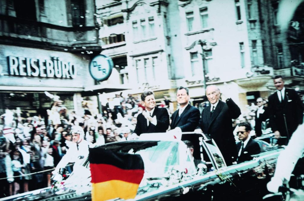 Kennedy-in-Berlin-1963, er steht mit Willy Brandt und Konrad Adenauer in einem Auto und fährt an einem DER Reisebüro vorbei