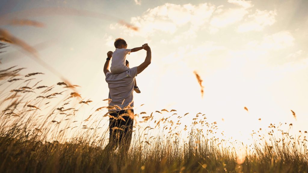 Rückansicht Vater_mit_Sohn_auf den Schultern in einem Feld, beide schauen in Richtung Sonne